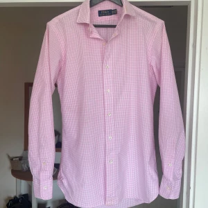 Rosa rutig skjorta från Ralph Lauren - Säljer en rosa och vit rutig skjorta från Ralph Lauren med klassisk krage och grönt broderat logotyp på sidan. Skjortan har långa ärmar och knäppning framtill. Perfekt för dig som gillar stilrena och tidlösa plagg. Sjukt fräsch till sommaren med ett par feta shorts! OBS! Har fläck vid kragen och längst ned vid knapparna, men man ser de knappt, se bild.    