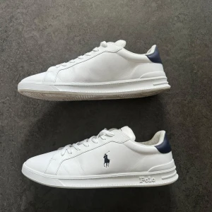 Vita sneakers från Polo Ralph Lauren - Säljer ett par fräscha vita sneakers från Polo Ralph Lauren med mörkblå detalj vid hälen och klassisk logga på sidan. Skorna har snörning och rund tå, perfekt för en stilren look. Kommer med ett par polo snören också.