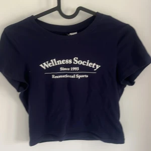 Mörkblå croppad t-shirt från Divided - Säljer en mörkblå croppad t-shirt från Divided med vit text 'Wellness Society' framtill. T-shirten har rund halsringning och korta ärmar. Perfekt för en avslappnad stil.