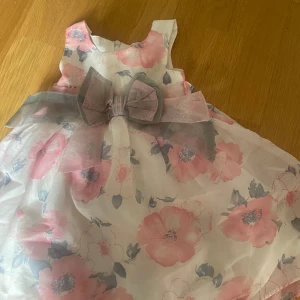Blommig klänning med rosett från Little Aimy - Supersöt blommig klänning från Little Aimy i vitt med stora rosa och grå blommor. Storlek 98-104