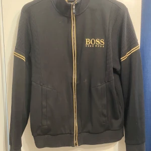 Svart jacka från Hugo Boss - Snygg svart jacka från Hugo Boss med gulddetaljer och dragkedja. Jackan har långa ärmar och ribbade muddar. Perfekt för en stilren look. Storlek S