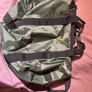 Grön axelväska med svart rem - Snygg grön axelväska med svarta remmar och en gul logga. Perfekt för vardagsbruk med gott om utrymme. Tillverkad i slitstarkt syntetmaterial.