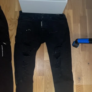 Dsquared2 svarta - Säljer ett par svarta jeans med coola slitningar och färgstänk. De har en dragkedja och knapp framtill. Perfekta för en edgy look!