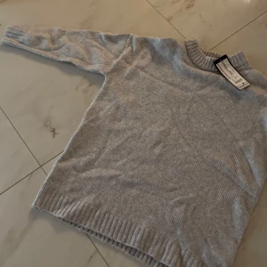 Grå stickad tröja - Säljer en mysig grå stickad tröja med rund hals och långa ärmar. Perfekt för kyliga dagar med ribbade muddar vid ärmslut och nederkant. Enkelt och stilrent plagg för garderoben. Har lappen och nytt skick