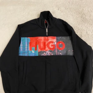 Svart tröja med tryck från Hugo - Snygg svart tröja från Hugo med ett färgglatt tryck och logotyp på framsidan. Tröjan har en dragkedja vid halsen och långa ärmar. Perfekt för en stilren look.