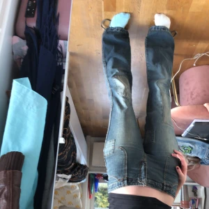 jeans med patchar från Jennyfer - Säljer ett par coola blå jeans från Jennyfer med unika patchar och slitna detaljer. Byxorna är bootcut. De har fickor både fram och bak samt justerbara snören vid bensluten. De är midrise. Ena bakfickan saknar en knapp.