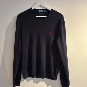 Svart tröja från Polo Ralph Lauren - Säljer en stilren marinblå tröja från Polo Ralph Lauren i 100% Pima bomull. Tröjan har en klassisk v-ringning och ett rött broderat märke på bröstet. Perfekt för en elegant och avslappnad look.