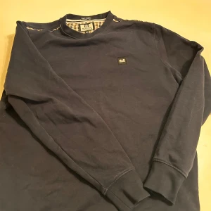 Mörkblå tröja från Weekend Offender - Säljer en stilren mörkblå tröja från Weekend Offender. Tröjan har långa ärmar och en rund halsringning med ett rutigt mönster på insidan av kragen. Perfekt för du som vill ha en casual tröja. Aldrig andvänd bara testad, köpt från weekend offenders egna hemsida. 