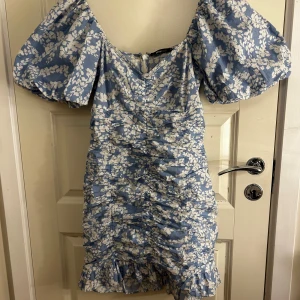 Blå blommig klänning från Gina Tricot - Säljer en söt blå klänning med vita blommor från Gina Tricot. Klänningen har puffiga kortärmar och en volangdetalj vid fållen. Den har en dragkedja i ryggen och en smickrande passform med rynkade detaljer. Perfekt för sommarens alla tillfällen! 🌸