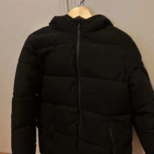 Svart pufferjacka från Supply & Demand - Säljer en svart pufferjacka från Supply & Demand med huva och dragkedja. Jackan har en cool logga på ärmen och är perfekt för kyligare väder. Den är designad för att hålla dig varm och bekväm.