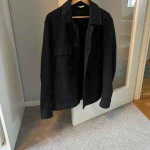 Svart overshirt med knappar - Snygg svart overshirt med knappar och stora fickor framtill. Perfekt för lager-på-lager-stilen. Den har långa ärmar och en klassisk krage. En stilren och mångsidig jacka för olika tillfällen.  Riktigt bra skick men säljer nu denna eftersom den är för liten för mig! 