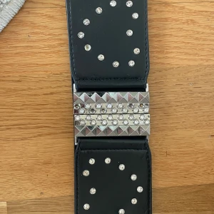 Svart bälte med nitar och strass - Snyggt svart bälte dekorerat med glittrande strass och silverfärgade nitar. Bältet har en bred design med en iögonfallande spänne i silver. Perfekt för att ge en edgy touch till din outfit.
