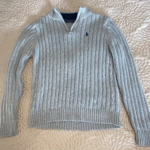 Grå stickad tröja från Ralph Lauren - Säljer en stilren grå stickad tröja från Ralph Lauren med kabelstickat mönster och en broderad logga på bröstet. Tröjan har en dragkedja vid halsen och långa ärmar. Perfekt för kyliga dagar!