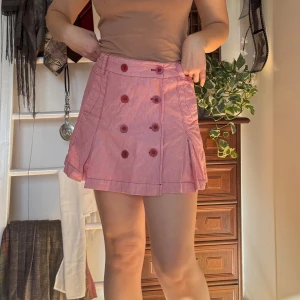 Rosa kortkjol med knappar - Köpt secondhand och använts en del. Bra skick. Säljer en söt rosa kortkjol med dubbla rader av knappar framtill. Kjolen har en A-linjeform och dekorativa fickor bak. Perfekt för en somrig look!