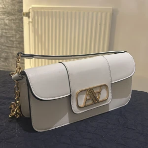 Vit axelväska med guldkedja - Snygg vit axelväska med elegant guldkedja och stilren design. Väskan har ett dekorativt guldfärgat spänne på framsidan och en praktisk axelrem. Perfekt för att ge din outfit en lyxig touch.