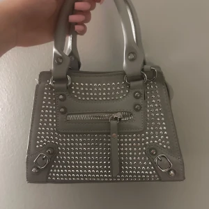 Grå handväska med nitar - Snygg och trendig handväska i silver med glittriga nitar och dragkedja framtill. Man kan knipsa på ett justerbart band så att man kan ha väskan hängandes från axeln. Perfekt för att ge din outfit en extra touch. 