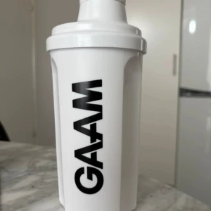 Vit shaker från GAAM - Säljer en vit shaker från GAAM med svart logotyp. Shakern har ett avtagbart lock och en insats med bikakemönster för effektiv blandning. Perfekt för proteinshakes och andra drycker. Enkel att rengöra och praktisk att använda både hemma och på gymmet. Aldrig använd!