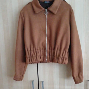 Brun jacka från Ambar - Snygg brun jacka från Ambar med dragkedja framtill och en klassisk krage. Jackan har en elastisk midja för en bekväm passform och långa ärmar. Perfekt för höstvädret!