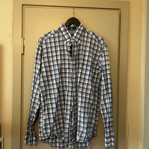 Blå och vit rutig skjorta från GANT - Säljer en klassisk blå och vit rutig skjorta från GANT i storlek XL. Skjortan är tillverkad i 100% bomull och har långa ärmar med knappar framtill. Perfekt för en stilren look.