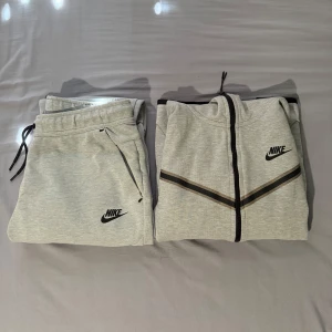 Gråa Nike tracksuit - Säljer en grå träningsjacka och matchande byxor från Nike. Jackan har dragkedja och huva, med svarta detaljer och Nike-logga på bröstet. Byxorna har dragsko i midjan och sidofickor med dragkedja. Perfekt för träning eller en avslappnad dag. Det är storlek S på hoodien och M på byxorna. Pris kan diskuteras