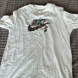 Vit t-shirt från Nike med färgglatt tryck - Säljer en vit t-shirt från Nike med ett coolt och färgglatt tryck på framsidan. T-shirten har en normal passform och korta ärmar. Perfekt för en avslappnad stil. Skicka ett meddelande vid intresse!