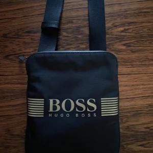 Hugo Boss Axelremsväska - Helt ny Hugo Boss Axelremsväska helt fel fri. Den har två stycken fack och ett band som går att spänna eller lösa beroende på vilken längd man vill ha. One size