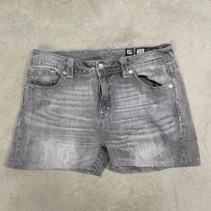 Grå jeansshorts från Miss Me - Snygga grå jeansshorts från Miss Me med slitna detaljer och broderad logga på bakfickan.                                                            Midjemått tvärs över:  41cm                                  Längd: 32 cm 