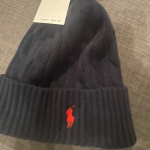 Mörkblå ribbad mössa från Ralph Lauren - Snygg mörkblå mössa från Ralph Lauren med ribbad design och röd broderad logotyp framtill. Perfekt för att hålla värmen under kyliga dagar.