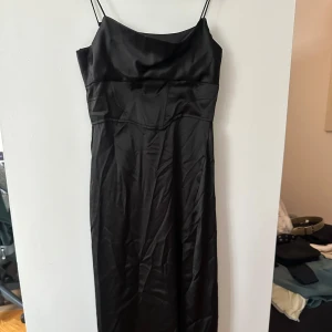 Svart lång axelbandslös klänning från Zara - Elegant svart klänning från Zara med smala axelband och en stilren design. Klänningen är tillverkad i ett glansigt material som ger en lyxig känsla. Perfekt för en kväll ute eller en speciell tillställning. Sitter likt en korsett vid midjan.