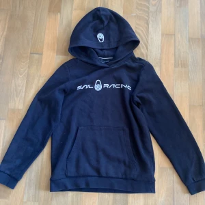 Svart hoodie från Sail Racing - Säljer en svart hoodie från Sail Racing med tryck på bröstet och en praktisk magficka. Hoodien har en justerbar huva och långa ärmar, perfekt för kyliga dagar. Passar både till vardags och för en sportig look. 🖤