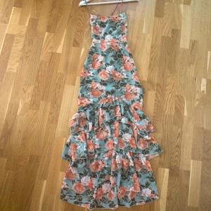 Blommig långklänning med volanger - Aldrig använd! Säljer en supervacker långklänning med blommigt mönster i fina vår/sommarfärger. Klänningen har smala axelband med knytning i bak och flera lager av volanger nertill som ger en romantisk känsla. Perfekt för bröllop och andra sommar festligheter!