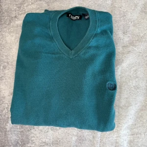 Grön v-ringad tröja från Chaps - Säljer en snygg grön v-ringad tröja från Chaps i 100% bomull. Tröjan har långa ärmar och en diskret logga på bröstet. Perfekt för en stilren look.