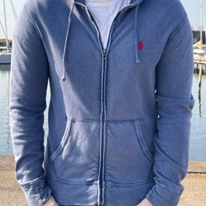 Blå hoodie från Ralph Lauren - Säljer en snygg blå hoodie från Ralph Lauren med dragkedja och det klassiska röda logotypen på bröstet. Tröjan har långa ärmar och en bekväm passform, perfekt för kyliga dagar. Den har också en praktisk huva med dragsko. De är storlek S men den passar bättre som M