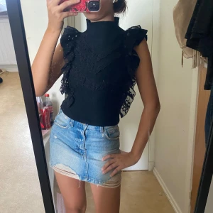 Svart Zara volang topp med spetsdetaljer - Nysick Supersnygg svart topp med hög hals och volanger längs axlarna. Toppen är ärmlös och har spetsdetaljer framtill i ett V-mönster. Materialet är stickat och stretchigt, vilket gör den både bekväm och trendig. Perfekt för dig som vill sticka ut lite extra.