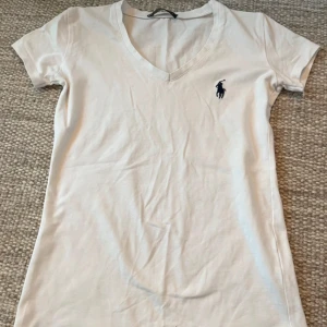 Vit v-ringad t-shirt från Ralph Lauren - Vit t-shirt från Ralph Lauren med v-ringning och korta ärmar. Klassisk design med mörkblå logga broderad på bröstet. Skön och mjuk bomull som passar perfekt till jeans eller shorts.