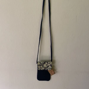 Mörkblå mobilväska med blommigt lock - Snygg liten axelväska att förvara sin mobil i. Mörkblå bomull med långt axelband. Locket har ett blommigt mönster i beige och stängs med en silverfärgad knapp. Perfekt för mobilen och småprylar när du vill ha händerna fria.