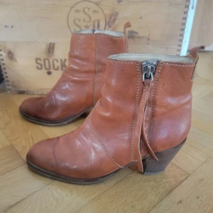 Bruna boots i skinn med klack - Snygga bruna boots i äkta skinn med dragkedja på sidan och dekorativ rem. Rund tå och stabil klack ger en cool och avslappnad vibe. Perfekta till jeans eller klänning för en trendig look.