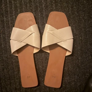 Beige sandaler med korsade remmar - Stilrena beige sandaler från H&M med breda korsade remmar i syntetmaterial. De har fyrkantig tå och platt sula, perfekt för en trendig look under varma dagar.