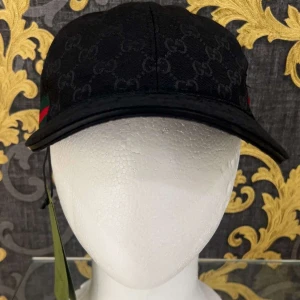 Svart keps från Gucci - Svart keps från Gucci med diskret GG-monogram över hela kepsen och klassiska gröna och röda ränder på sidan. Justerbar rem baktill i skinn och böjd skärm. Snygg och stilren accessoar som lyfter din outfit.