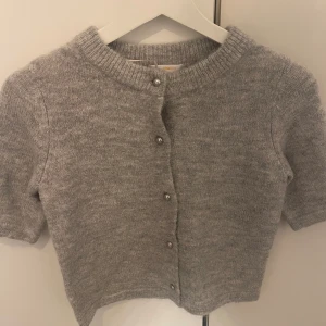 Grå stickad kofta med kort ärm - Superfin grå stickad kofta med korta ärmar och rund hals. Koftan har snygga knappar framtill och ribbade kanter vid hals och ärmslut. Perfekt att slänga över en topp för en chill och stilren look.
