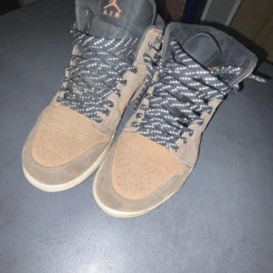 Nike Air Jordan 1 High Travis Scott - Säljer ett par Nike Air Jordan 1 High Travis Scott med bruna och beige mockadetaljer, svart swoosh och svarta/vita snören. Skorna har hög siluett och klassisk Jordan-logga på sidan. Perfekt för dig som gillar streetwear och unika sneakers. SKO LÅDA INGÅR