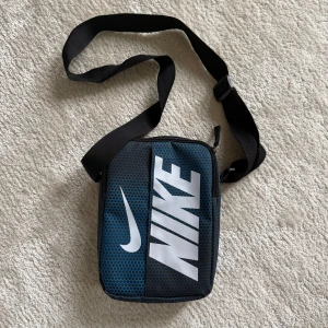 Nike axelväska med blått hexagonmönster - Snygg axelväska från Nike i blått och svart med hexagonmönster och stor vit logga framtill. Väskan har justerbar axelrem, dragkedja upptill och extra fack med dragkedja på baksidan. Perfekt storlek för mobil, plånbok och småprylar.
