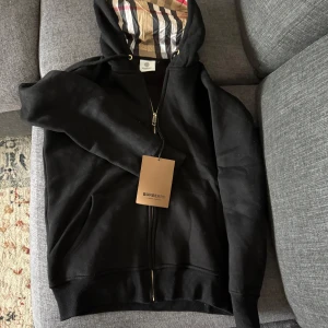 Svart hoodiejacka från Burberry - En helt ny Svart hoodiejacka från Burberry med dragkedja och klassisk rutig huva i beige, svart, vit och röd. Jackan har snörning i huvan och ribbade muddar. Perfekt för dig som vill ha en stilren och lyxig streetwear-look.