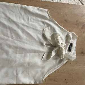 Vit ärmlös blus med knyt från Sisters Point - Stilren vit blus från Sisters Point i ärmlös modell. Toppen har en snygg knytning vid halsen som ger en chic touch. Tillverkad i ett lätt och mjukt material som faller fint. Bra att ha under cardigans så att  rosetten syns - utan knöggliga armar.