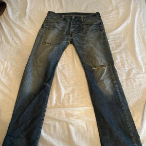 Levi's 501 slitna blå jeans W33 L34 - Klassiska Levi's 501 jeans i blå tvätt med slitningar och hål på båda benen för en cool, avslappnad look. Rak passform, fem fickor och knappgylf. Perfekta för dig som gillar vintage vibe och streetstyle. w33 L34. PRISFÖRSLAG
