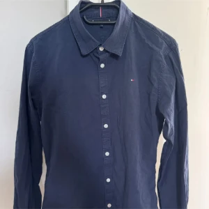 Mörkblå skjorta från Tommy Hilfiger - Klassisk mörkblå skjorta från Tommy Hilfiger med vit knappstängning och diskret logga broderad på bröstet. Skjortan har lång ärm, normal passform och är tillverkad i mjuk bomull. Perfekt till jeans eller chinos för en clean look.