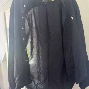  Polo Ralph Lauren - Snygg svart skjorta från Polo Ralph Lauren i klassisk passform. Tillverkad i tunn och luftig linne, perfekt för varmare dagar. Skjortan har långa ärmar, knappar framtill och den ikoniska broderade loggan på bröstet.