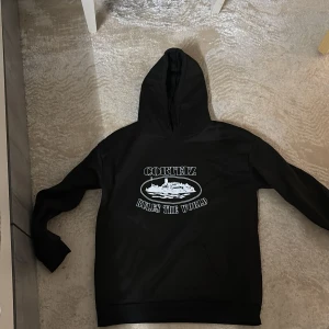 Svart Corteiz hoodie med tryck - Svart hoodie från Corteiz med vit text och motiv på bröstet där det står 'Corteiz Rules The World'. Klassisk huva och känguruficka framtill. Perfekt för dig som gillar streetwear och vill sticka ut med en snygg logga.