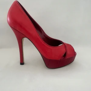 Röda platåpumps från Via Uno - Snygga röda pumps med hög klack och platåsula från Via Uno. Storlekar 35-41. Skorna har öppen tå och korslagda remmar framtill. Tillverkade i skinn med en elegant, spetsig siluett som verkligen sticker ut.