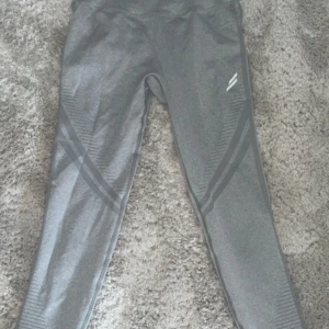 Grå leggings från DoYouEven - Snygga grå leggings från DoYouEven med hög midja och scrunch-detalj bak. De har ribbade partier och diskreta ränder längs benen för en sportig look. Perfekta för gymmet eller chill hemma. Materialet är stretchigt och följsamt för bästa komfort.